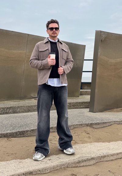 Camisa de cuadros marrón sobre un suéter negro, con una camisa de rayas azul claro debajo. Jeans oscuros y zapatillas plateadas, sosteniendo una taza.