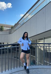 Camicia azzurro chiaro con bottoni, pantaloni corti neri con strisce laterali, scarpe nere e calzini bianchi. In piedi su un railing in un contesto urbano.