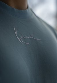 Mörkblå bomullströja med rund hals; har en ljusfärgad broderad signatur på bröstområdet. Nära vy framhäver den mjuka texturen.