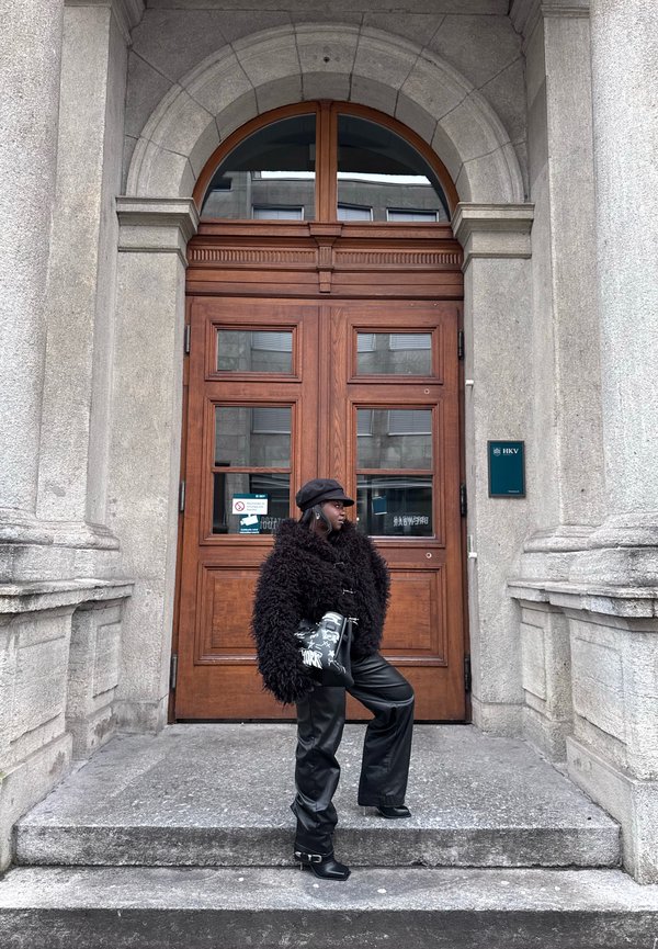 Femeie într-o jachetă neagră din blană artificială, pantaloni de piele și pălărie, stând pe trepte de piatră și ținând o geantă neagră cu model în fața unor uși duble mari din lemn.