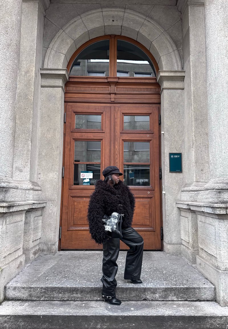 Femme vêtue d'une veste en fausse fourrure noire, d'un pantalon en cuir et d'un chapeau, debout sur des marches en pierre, tenant un sac noir à motifs devant de grandes portes doubles en bois.