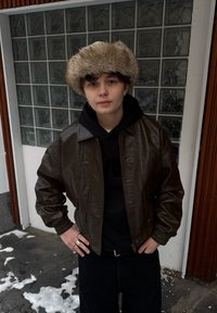 Veste aviateur en cuir marron avec poignets élastiques, portée par-dessus un sweat à capuche noir. Porte un chapeau en fourrure légère et duveteuse de couleur claire. Neige et trottoir visibles.