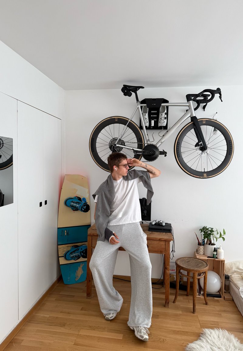 Personne en pantalon de survêtement gris et chemise blanche pose à l’intérieur près d’un bureau en bois, d’un vélo accroché au mur, d’un skateboard et de petits tabourets avec des plantes.