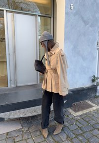 Beżowy płaszcz trencz z paskiem w talii i pagonami na ramionach, zestawiony z czarnymi spodniami o szerokich nogawkach i szarą dzianinową czapką. Czarna pleciona torba i taupe botki za kostkę.