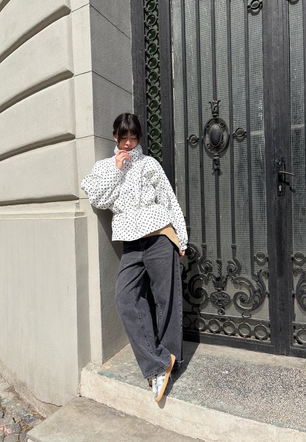 Persoană tânără sprijinită de un perete în colț, purtând o geacă albă cu buline, blugi gri cu picioare largi și adidași lângă o poartă decorativă din metal negru.