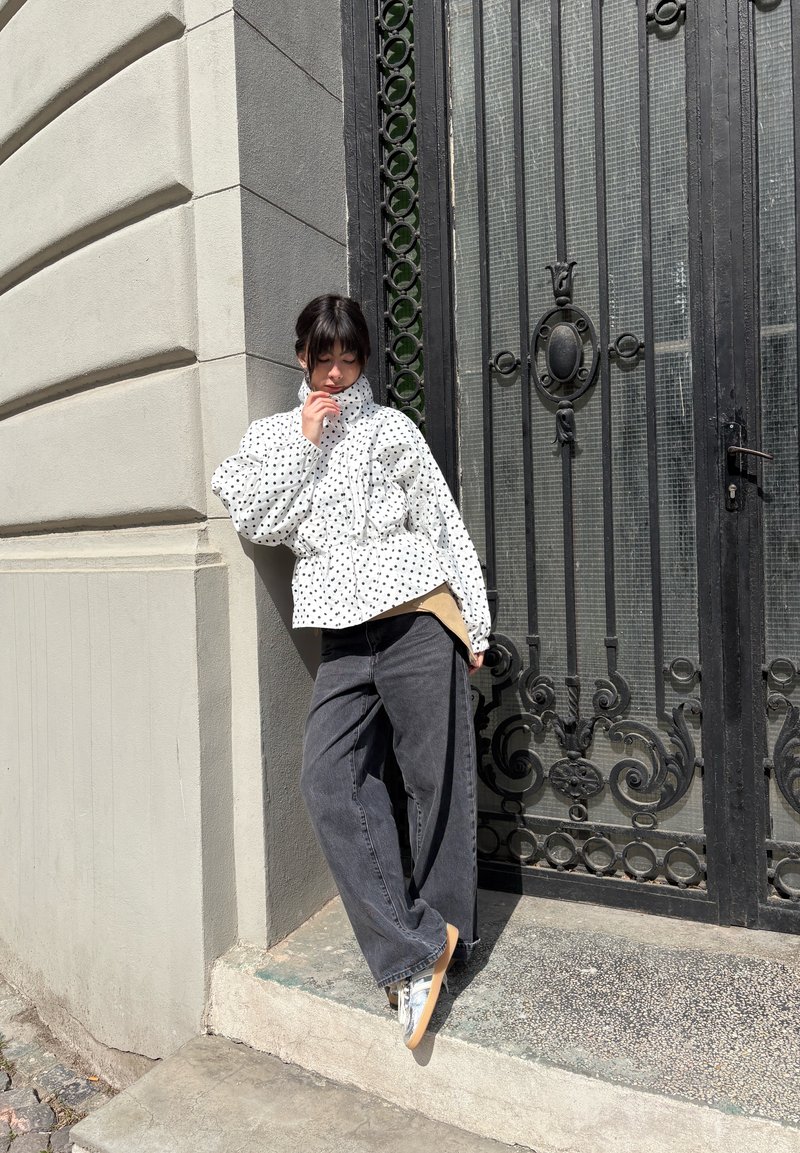 Jeune personne appuyée contre un mur d'angle, portant une veste blanche à pois, un jean gris à jambes larges et des baskets près d'une grille décorative en métal noir.