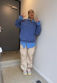 Avslappet antrekk: blå oversized genser over en lyseblå skjorte med knapper, beige cargo-bukser og flerfargede joggesko. Innendørs setting med en dør.