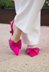 Forró rózsaszín slingback magassarkú cipők, hegyes orral, nagy, csillogó masnikkal díszítve, különböző rózsaszín árnyalatokban, világos színű felületen elhelyezve.
