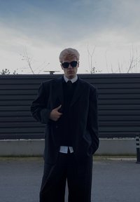 Jeune homme aux cheveux blonds portant des lunettes de soleil noires, un blazer noir surdimensionné, une cravate et un pantalon à carreaux, debout en plein air au crépuscule.