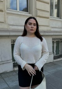 Pull en maille beige clair à manches longues, associé à une jupe noire. Un sac crème est porté, avec un fond texturé d'un bâtiment.