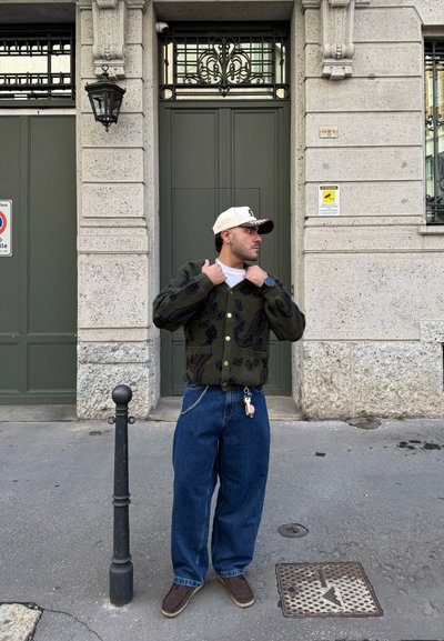 Cárdigan verde oscuro con cierre de botones, diseño estampado, combinado con jeans azules holgados y zapatos marrones. De pie junto a un poste negro.