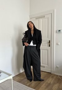 Black oversized coat with fur trim, black cardigan, white vest, and loose grey trousers. Black pointed shoes and a dark handbag.