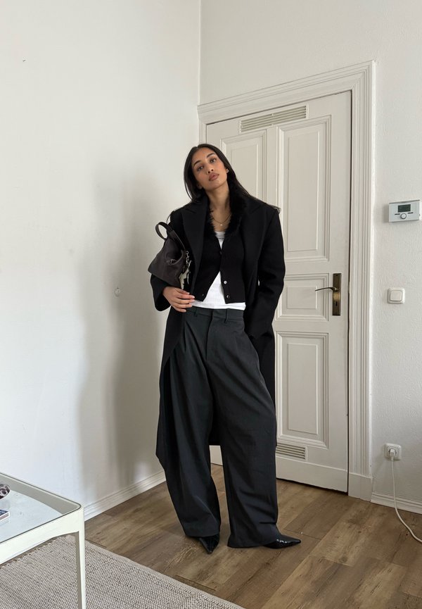 Manteau noir surdimensionné avec garniture en fourrure, cardigan noir, débardeur blanc et pantalon gris ample. Chaussures noires pointues et sac à main sombre.