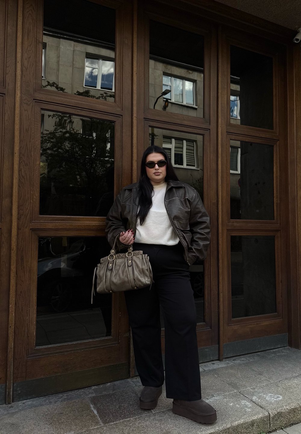 Giacca di pelle marrone sopra un maglione bianco, pantaloni larghi neri, stivali con platform marroni e una borsa grigia con hardware dorato. Ambientazione all'aperto.