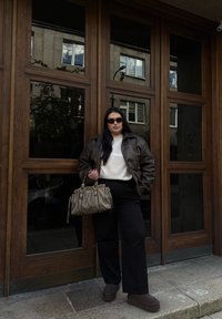 Giacca di pelle marrone sopra un maglione bianco, pantaloni larghi neri, stivali con platform marroni e una borsa grigia con hardware dorato. Ambientazione all'aperto.
