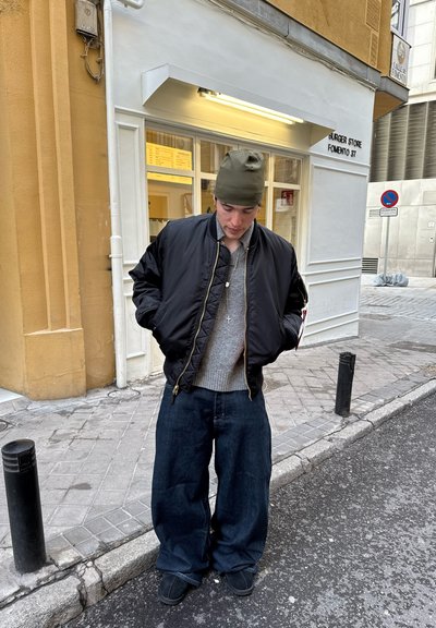 Chaqueta bomber negra, suéter gris, jeans oscuros de gran tamaño y un gorro verde. Está de pie en una calle de adoquines cerca de un restaurante.