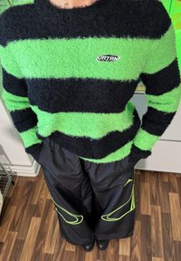 Svart og grønn stripete, fluffy genser med logo på brystet, kombinert med løse svarte bukser med grønne detaljer og søm.
