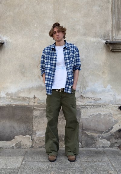 Joven con las manos en los bolsillos, viste una camisa azul a cuadros sobre una camiseta blanca, pantalones verde oliva y zapatos marrones, de pie frente a una pared envejecida.