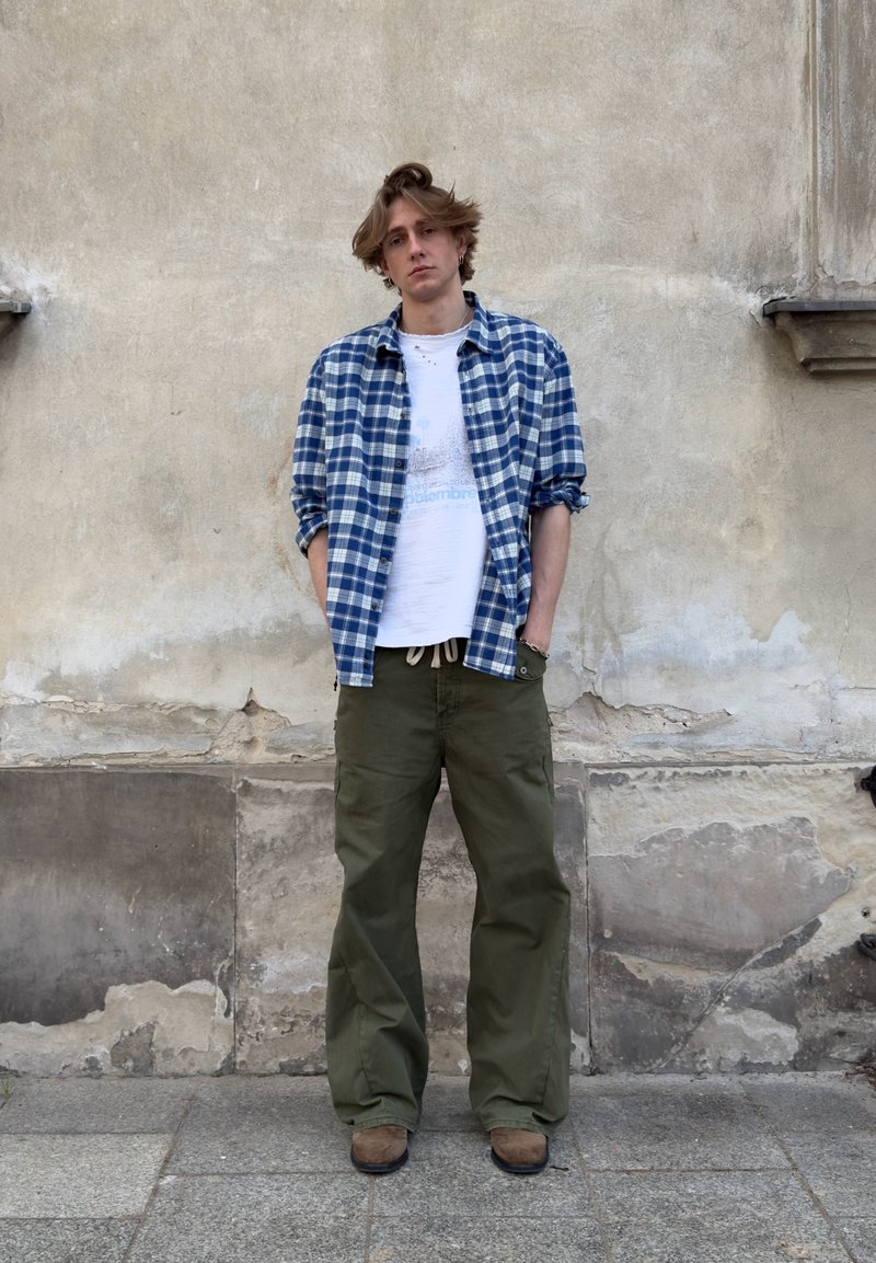 Jeune homme les mains dans les poches, portant une chemise à carreaux bleue par-dessus un t-shirt blanc, un pantalon vert olive et des chaussures marron, debout contre un mur abîmé.