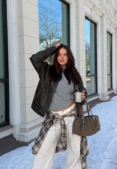 Mujer joven con ropa de invierno sosteniendo una taza de café y un bolso marrón, de pie en una acera nevada junto a un edificio con grandes ventanales.