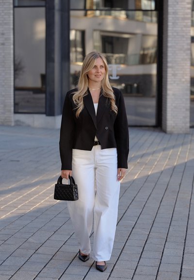 Mujer con cabello rubio largo camina por la acera con chaqueta negra, pantalones blancos, lleva un bolso negro acolchado, tacones negros, cerca de un edificio moderno.