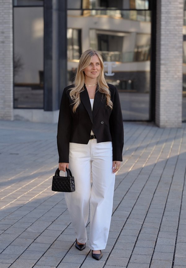 Woman with long blonde hair walks on pavement wearing black blazer, white trousers, holding black quilted handbag, black heels, near modern building.