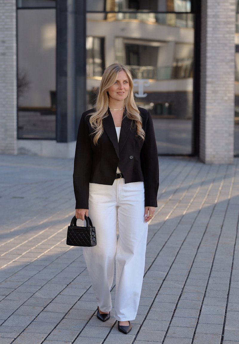 Femme aux longs cheveux blonds marche sur le trottoir portant un blazer noir, un pantalon blanc, tenant un sac à main noir matelassé, des talons noirs, près d'un bâtiment moderne.