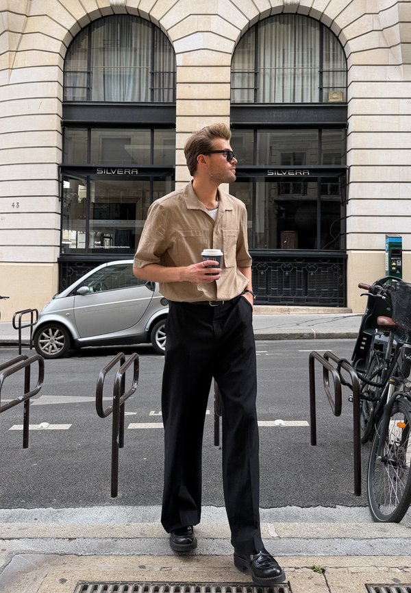 Hombre con gafas de sol y camisa beige sostiene una taza de café mientras está de pie cerca de los aparcamientos para bicicletas en una calle de la ciudad frente a un edificio.