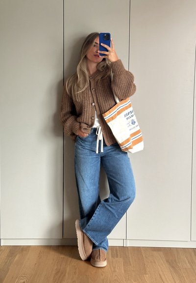 Cárdigan de punto marrón con botones, camiseta blanca, jeans de pata ancha azul claro y zapatos tipo slip-on beige. Lleva un bolso de lona a rayas.
