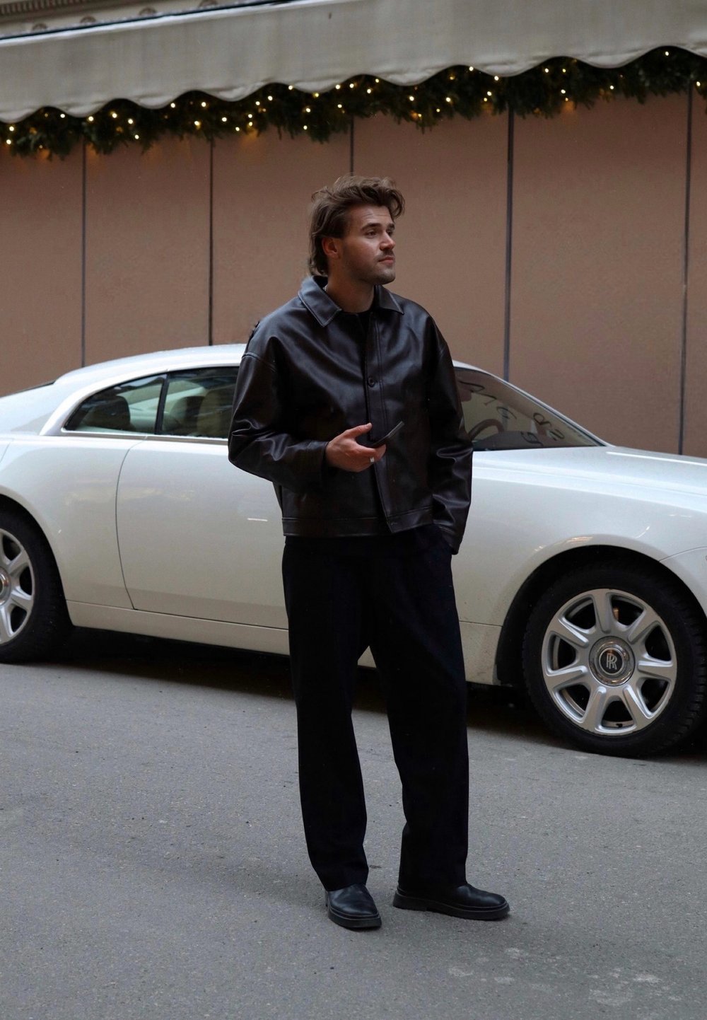 Chaqueta de cuero negra con cuello, combinada con pantalones oscuros y zapatos negros. Un coche de lujo blanco está aparcado al fondo.