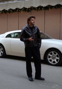Chaqueta de cuero negra con cuello, combinada con pantalones oscuros y zapatos negros. Un coche de lujo blanco está aparcado al fondo.