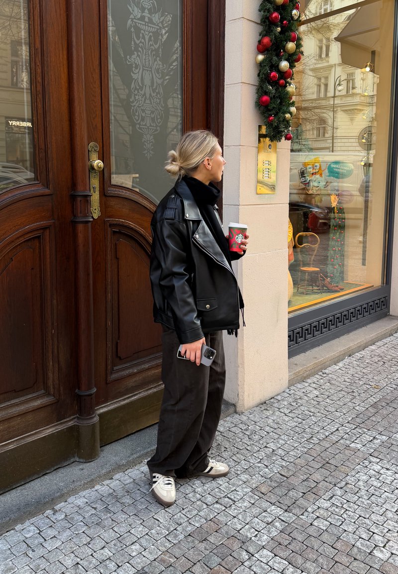 Frau in schwarzer Lederjacke, die Kaffee und Telefon hält, steht auf einer Kopfsteinpflasterstraße und schaut auf ein Schaufenster mit Weihnachtsdekoration.