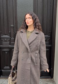 Bruine mantel van een wollen mix met een dubbele rij knopen en een gestructureerde afwerking. Voorzien van grote knopen, zakken en een reverskraag. Draagt een handtas met patroon.