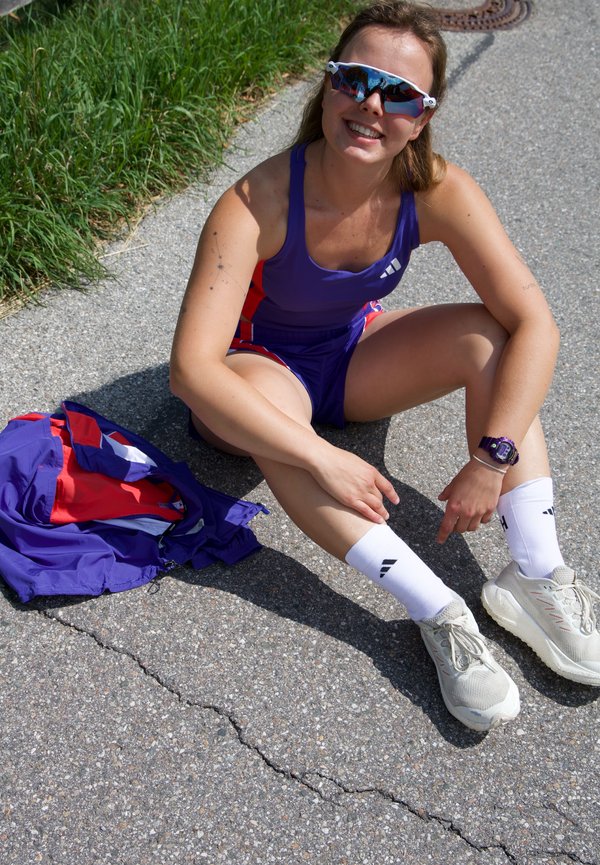 Kobieta w ubraniu sportowym i okularach przeciwsłonecznych siedzi na wybrukowanej ścieżce obok trawy z złożoną kurtką obok siebie, uśmiechając się do kamery.