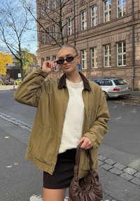 Prevelik jopič v olivno zeleni barvi z rjavim ovratnikom, bel teksturiran pulover, črna minikrilo in rjava torbica. Majhna črna sončna očala kot poudarek.