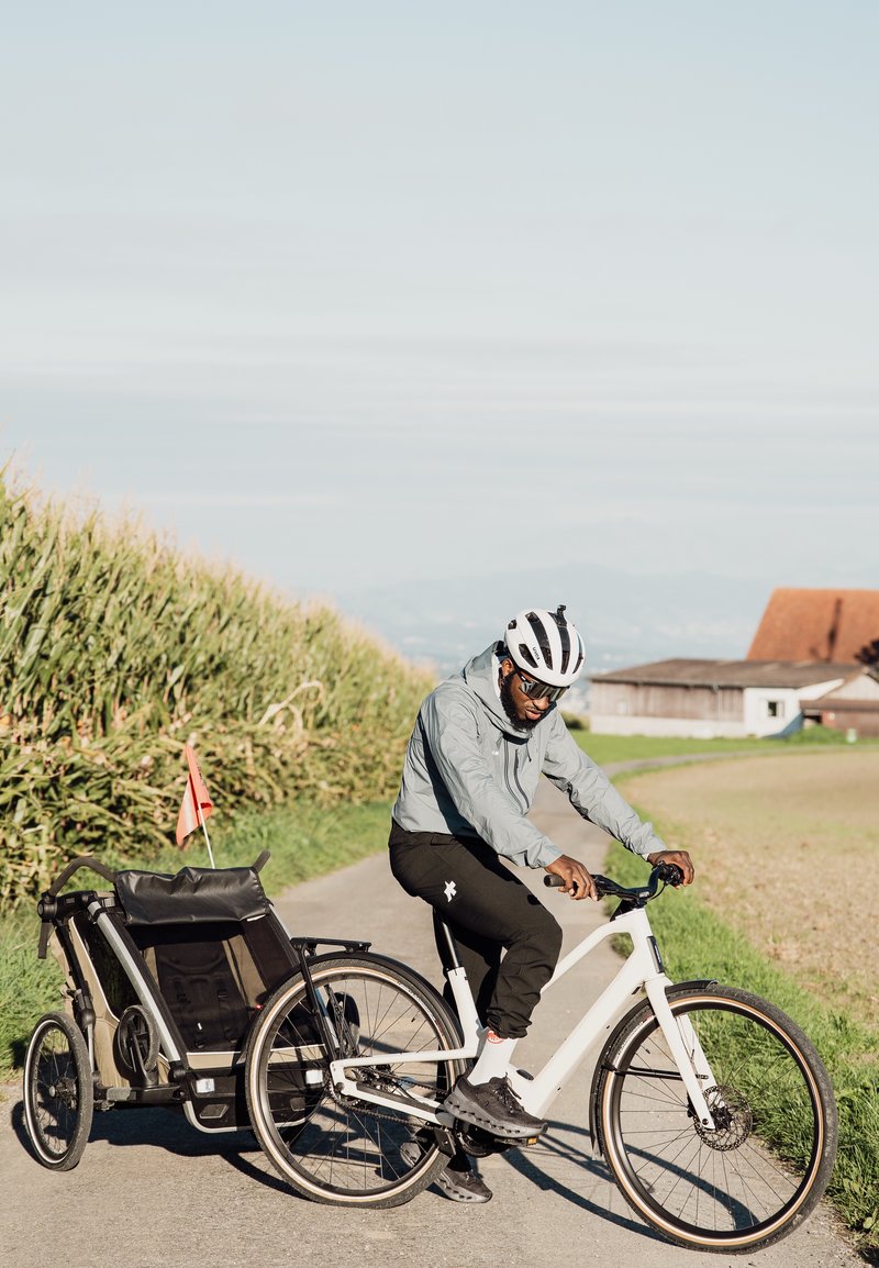 Vélo électrique avec un cadre blanc, des pneus noirs et une remorque de vélo en tissu noir. Le cycliste porte une veste grise et un casque, sur un chemin.