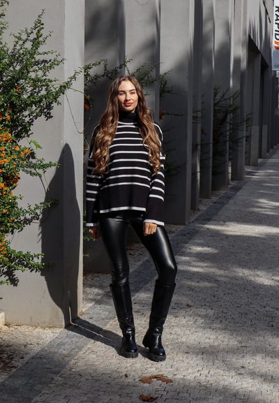 Suéter de cuello alto a rayas en blanco y negro, leggings de imitación cuero negro y botas negras hasta la rodilla. De pie en un entorno exterior brillante.