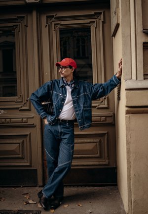 Hombre con gorra roja, gafas, chaqueta de mezclilla y vaqueros, apoyado contra una puerta de madera ornamentada y una pared beige.