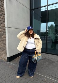 Beżowa kurtka sherpa, biała koszulka, ciemnoniebieskie dżinsy o szerokich nogawkach, granatowa czapka z logo, trzymając niebieską torebkę w wzory, stojąc na zewnątrz.