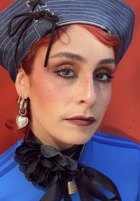 Persona con capelli rossi che indossa un cappello blu a righe, un orecchino a forma di cuore, rossetto scuro e un accessorio floreale nero intorno al collo, su uno sfondo rosso.