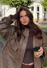 Brązowa, skórzana, oversize'owa kurtka z dużymi kieszeniami, noszona na ciemnej bluzce. Osoba trzyma kubek kawy i ma długie, ciemne włosy.