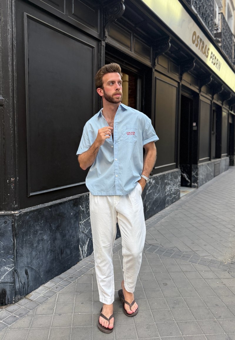 Homme portant une chemise bleu clair à manches courtes, un pantalon blanc et des tongs, marchant sur un trottoir en ville à côté d'une vitrine noire et en marbre.