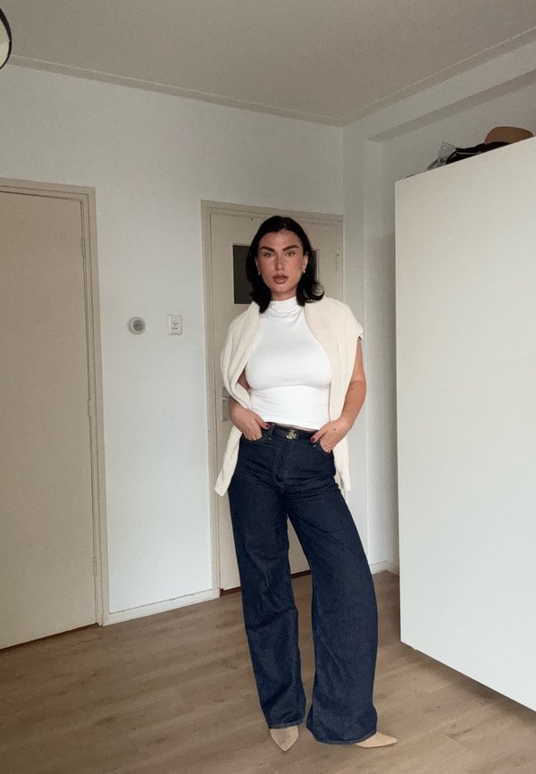 Mujer de pie en el interior, vestida con una camiseta blanca sin mangas, chaleco de punto color crema, jeans oscuros de pierna ancha y zapatos beige puntiagudos.