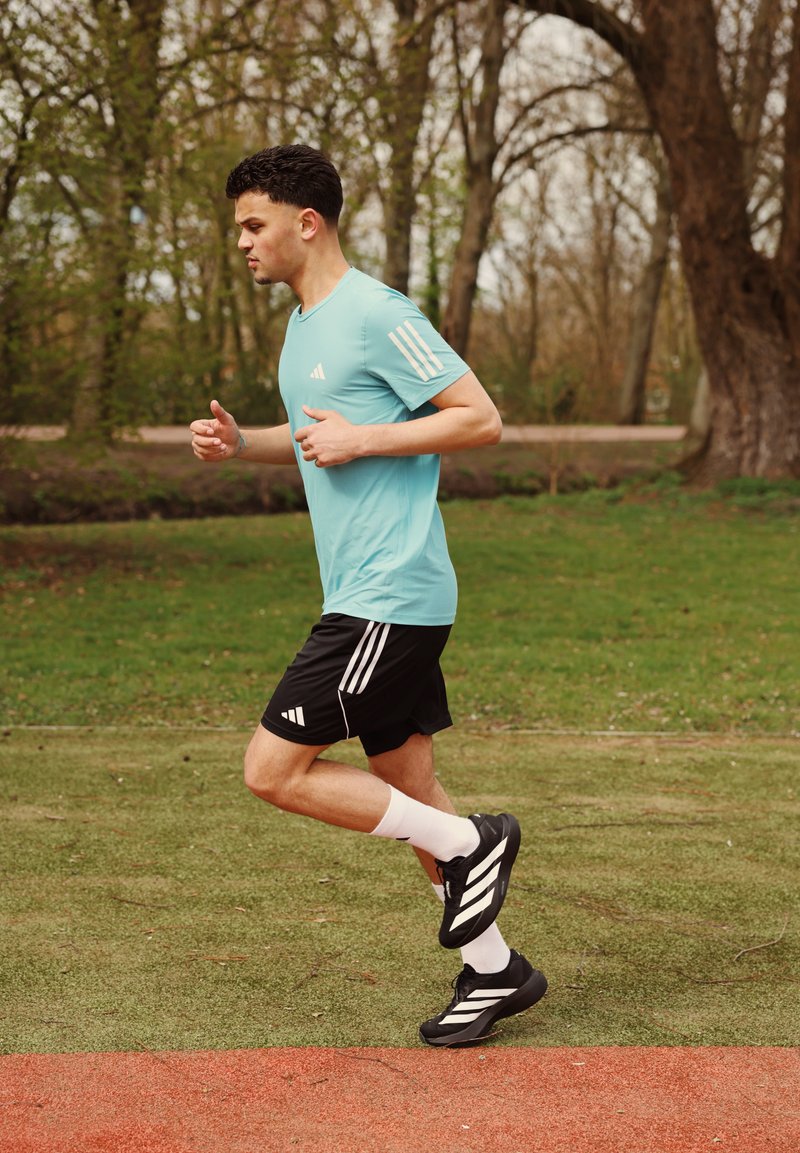 Homme faisant du jogging sur de l'herbe verte près d'une piste de course rouge, portant un t-shirt bleu clair, un short noir, des chaussettes blanches et des chaussures de course noires.