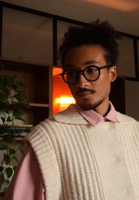 Pull en tricot de couleur claire avec une texture côtelée et un large col, porté par-dessus une chemise rose. L'arrière-plan présente des étagères en bois et de la verdure.