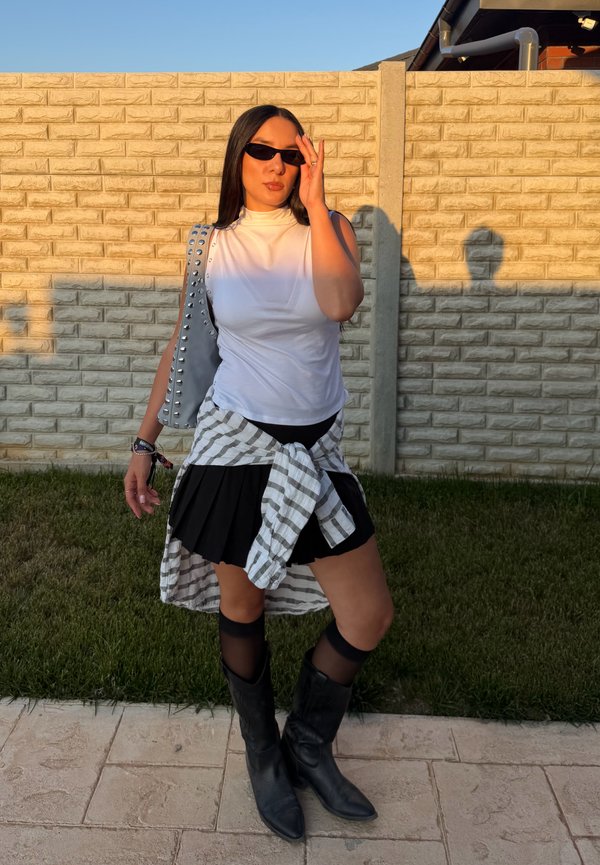 Femme portant des lunettes de soleil, un top blanc sans manches, une jupe noire, une chemise à carreaux nouée à la taille et des bottes noires, debout sur un trottoir près d'un mur en briques au coucher du soleil.