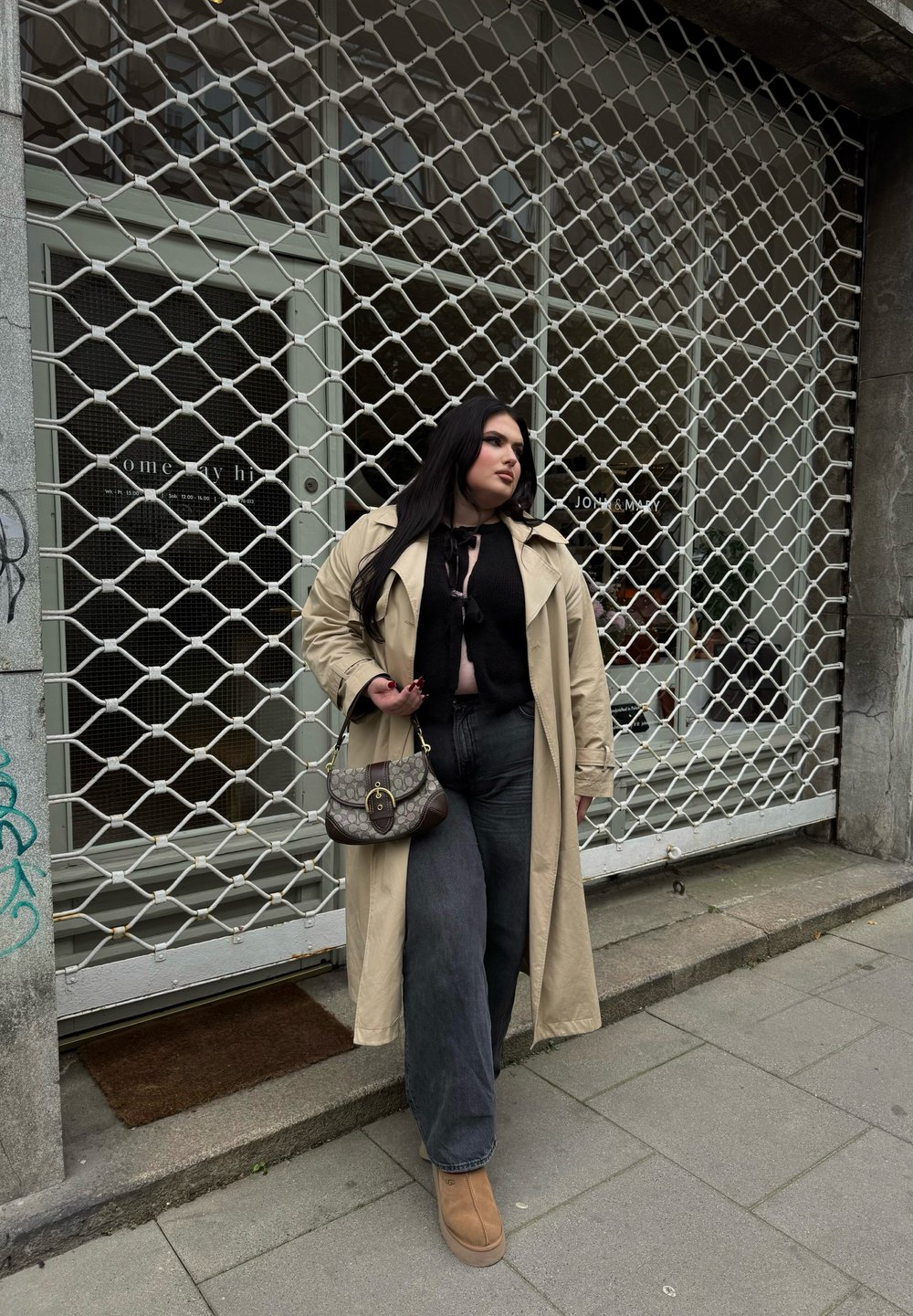 Bež plašč čez črn top, v kombinaciji z širokimi temnimi kavbojkami in rjavimi škornji. Drži rjav vzorčen ročaj, stoji ob trgovinskih vratih.