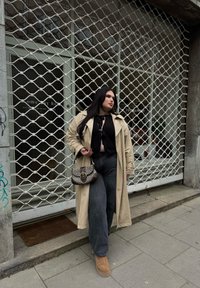 Bež plašč čez črn top, v kombinaciji z širokimi temnimi kavbojkami in rjavimi škornji. Drži rjav vzorčen ročaj, stoji ob trgovinskih vratih.