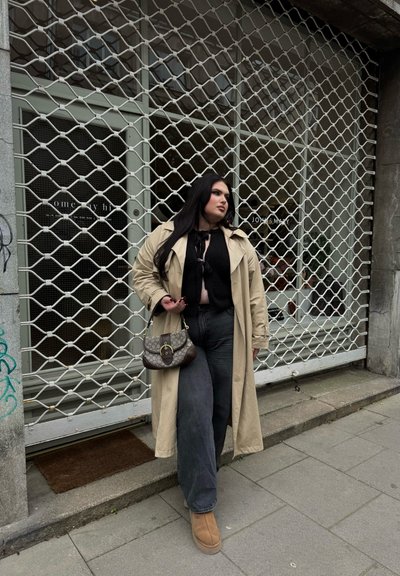 Gabardina beige sobre una blusa negra, combinada con unos vaqueros oscuros de pierna ancha y botas color topo. Sosteniendo un bolso de mano marrón con estampado, de pie junto a la puerta de una tienda.