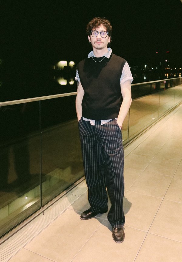 Ung mand med briller står på natlig terrasse iført sort ærmeløs sweater, stribede bukser og lædersko med hænderne i lommerne.